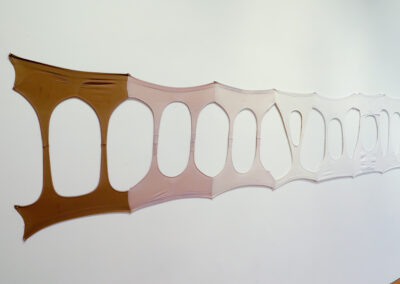 A series of nylon-like fabric pieces by Kailani Mayer in varying skin tones are stretched and attached to a white wall, forming an abstract, elongated pattern with oval cutouts.