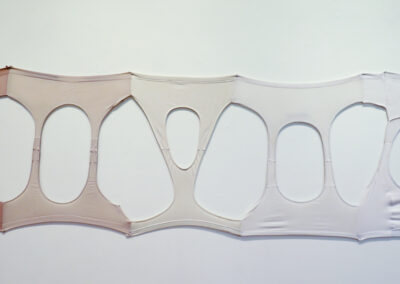 A row of light-colored pantyhose by Kailani Mayer is stretched horizontally on a white wall, with large oval and teardrop-shaped cutouts creating a repeating pattern. The material appears taut and slightly translucent.