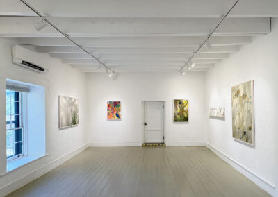 A bright art gallery room at El Zaguán with white walls, wooden floors, and a beamed ceiling displays colorful abstract paintings by Elizabeth Chiles. A single door is centered at the back wall, and a window lets in natural light on the left.