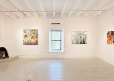 A bright, minimalist art gallery room with white walls and floors showcases four colorful paintings by Elizabeth Chiles. A small fireplace with stacked firewood sits in the corner, while a window fills the El Zaguán space with natural light.