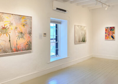 A bright, minimalist art gallery with white walls and a wooden floor at El Zaguán, displaying three colorful paintings by Elizabeth Chiles—sunflowers, abstract greenery, and bold shapes—bathed in natural light through a central window.
