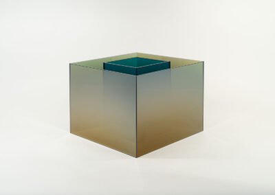 A translucent, gradient-colored glass cube with a smaller, similarly styled cube nested inside, both set against a plain white background. The Bell Mills 2025 design transitions from teal at the top to amber at the base.