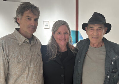 Three people pose and smile together indoors at Bell Mills. The woman in the center wears a black jacket, the man on the left wears a patterned shirt, and the older man on the right wears a hat and vest. Artworks are displayed in the background.