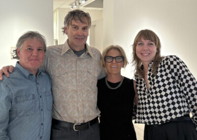 Four adults stand closely together indoors, smiling at the camera. The light-colored walls and ceiling lights hint at a gallery or exhibition space, possibly during a Bell Mills 2025 event.