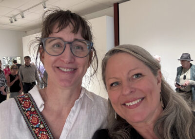 Two women smiling for a selfie at the Bell Mills 2025 art gallery, with people and artworks visible in the bright, modern background. One woman wears glasses; both look happy and relaxed.