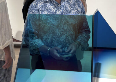 A smiling man in a blue patterned shirt stands behind a geometric glass sculpture with blue and green hues at Bell Mills 2025, while three people chat in the background.