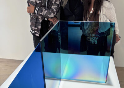 Three people stand smiling behind a modern geometric sculpture made of blue and iridescent glass panels, created for Bell Mills 2025, placed on a white pedestal in a gallery-like space.