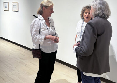 Three women stand and talk in the Bell Mills art gallery with white walls and wooden floors. Two framed artworks are visible behind them as they appear engaged in conversation, capturing the vibrant spirit of Bell Mills 2025.