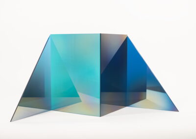 A geometric, angular sculpture made of translucent blue and turquoise panels, inspired by Bell Mills 2025, is arranged in a symmetrical, origami-like shape on a plain white background. Light creates subtle color gradients and reflections on the surfaces.