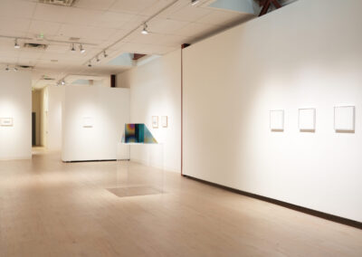 A contemporary art gallery with white walls, light wood floors, and minimalist artwork, including framed prints and a colorful geometric sculpture on a clear pedestal. The space is brightly lit with track lighting.