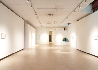 A spacious, brightly lit art gallery with white walls and light wood floors, displaying framed artwork and colorful pieces evenly spaced along the walls and a pedestal in the center.