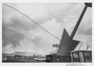 Black and white photo of a tilted, damaged structure with wires crossing the sky, a Kmart store in the background, and cloudy skies overhead; handwritten notes appear on the photo's lower right corner.