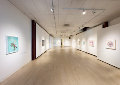 A spacious, well-lit art gallery with light wood floors and white walls displays colorful framed artwork by Munroe and McConville, evenly spaced along both sides of the hallway. The gallery appears empty of people, inviting quiet reflection in 2025.