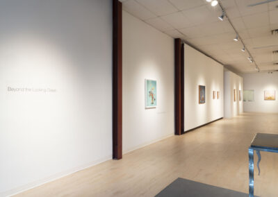 A bright, modern art gallery with light wood floors and white walls displays framed artwork. The wall on the left reads “Beyond the Looking Glass.” Track lighting illuminates the 2025 McConville Munroe exhibition along the corridor.