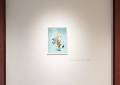 A framed painting of a colorful flower bouquet by McConville hangs on a white gallery wall under soft lighting. The name "Matthew McConville" and the year 2025 are displayed beside the artwork.