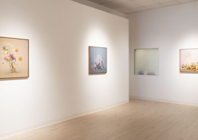 A minimalist art gallery room with light wood floors and white walls displays three colorful framed artworks by McConville Munroe, featuring abstract floral and balloon-like elements. A frosted glass window completes the 2025-inspired setting.