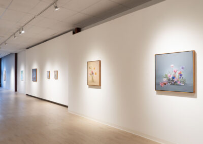 A bright art gallery hallway with light wooden floors and white walls, featuring McConville Munroe’s 2025 floral paintings spaced evenly and illuminated by ceiling spotlights.