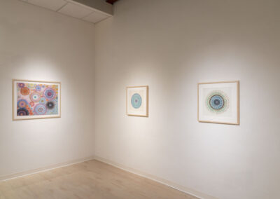 A gallery corner displays three framed abstract artworks on white walls; the left piece features colorful circular patterns, while the two on the right showcase mandala-like designs in blue and green tones by McConville Munroe for 2025.