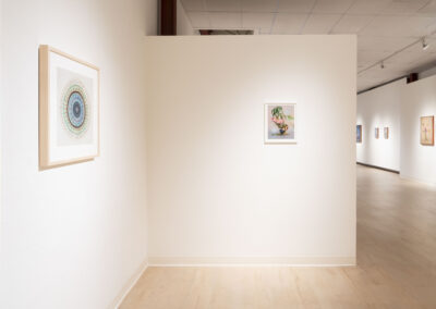 A bright art gallery features framed artworks on white walls, including a 2025 circular geometric piece on the left and a colorful McConville Munroe still life in the center, with additional art visible down the hallway.