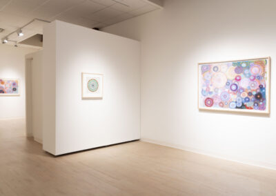 A bright art gallery with light wood floors and white walls, displaying three framed abstract artworks featuring colorful, circular patterns. The largest artwork by Munroe is on the right wall, with two smaller McConville pieces in the background.