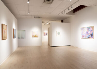 A bright, modern art gallery with light wood floors and white walls, displaying colorful abstract and floral paintings by McConville in wooden frames. Evenly spaced around the room, the artworks shine under ceiling lights, highlighting the 2025 collection.
