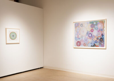 A gallery wall displays two framed abstract artworks by McConville Munroe featuring colorful concentric circles and patterns; one small piece hangs on the left, and a larger, more complex 2025 work is on the right, both against white walls and a wooden floor.