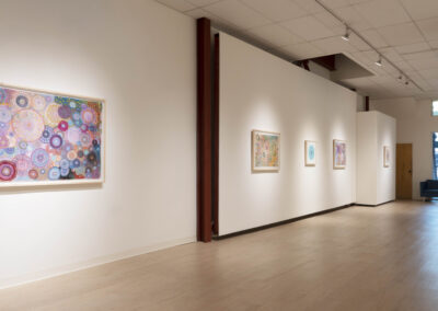 A modern art gallery with light wood floors and white walls displays colorful abstract paintings by McConville Munroe, primarily featuring circular patterns. Natural light enters from large windows in the background, reflecting a 2025 aesthetic.