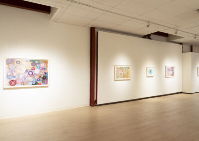 A bright art gallery with five colorful abstract paintings by McConville and Munroe on white walls, featuring circular and floral patterns. The space has wooden floors and a high white ceiling with spotlights, highlighting the 2025 collection.