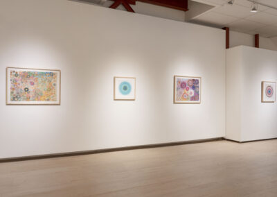 A gallery wall displays four framed abstract artworks by McConville Munroe, each featuring circular patterns and colorful designs, illuminated by spotlights, with a light wooden floor and white walls, perfect for a modern 2025 aesthetic.