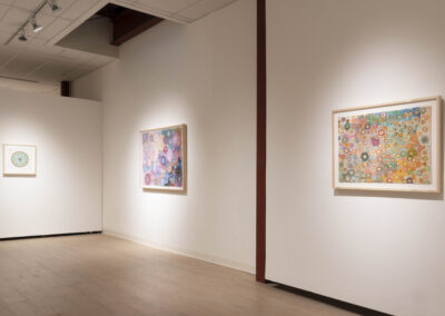 A gallery wall displays three framed abstract artworks by Munroe featuring colorful circular patterns and intricate designs. The room has light wooden floors and neutral walls, creating a bright, clean space for 2025.
