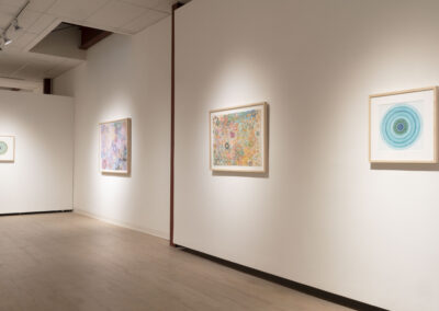 A gallery room with light wood floors and white walls, displaying four framed abstract artworks by Munroe featuring intricate, colorful patterns and mandala-like designs, evenly spaced along the walls for the 2025 season.