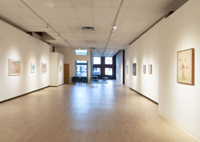 A bright, spacious art gallery with light wood floors and white walls displays colorful framed artworks, including pieces by McConville Munroe. Natural light enters through large windows at the far end, where seating is arranged for 2025 exhibitions.