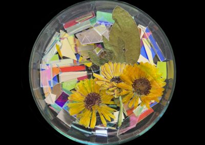A round glass dish contains three yellow dried flowers, two green leaves, and colorful shards of iridescent paper—an elegant display celebrating the Art of ReUse against a black background.