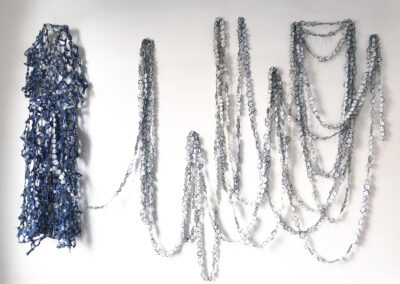 A wall installation of tangled, chain-like strands in blue and silver—crafted as stunning Upcycled Art—features a blue cluster on the left with silver drapes looping across the right, all set against a crisp white background.