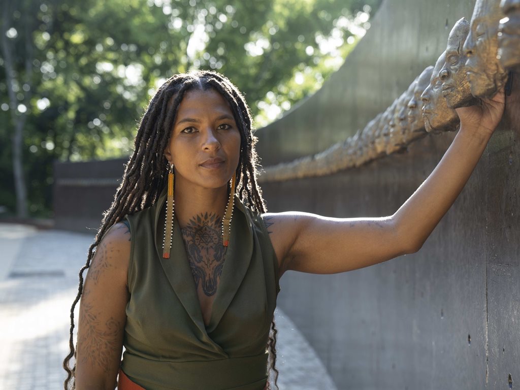 Portrait_Nikesha-Breeze_@Kate-Russell_Resized A woman with long braided hair and tattoos, possibly Nikesha Breeze, stands outdoors at the Biennale of Sydney, resting one arm on a curved bronze sculpture. She wears a sleeveless green top and long earrings. Trees and sunlight fill the background.