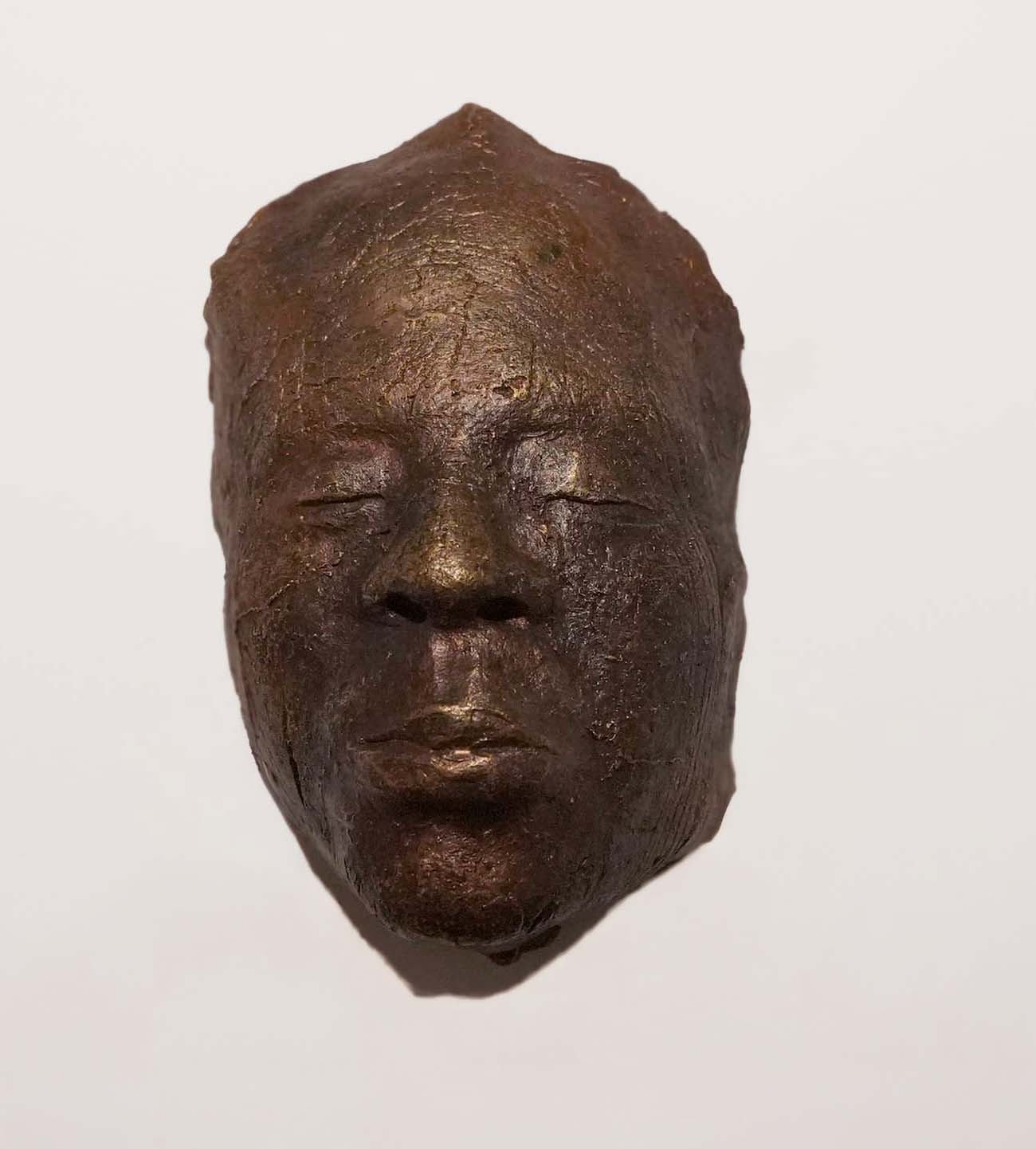 A bronze-colored sculpted mask of a human face with closed eyes and pursed lips, set against a plain white background.