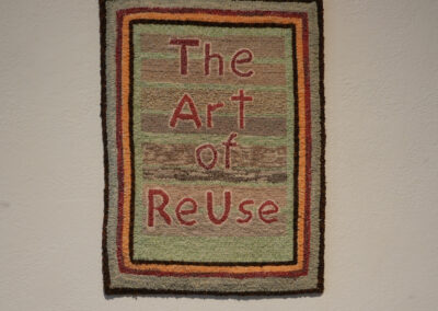A rectangular textile wall hanging with "The Art of ReUse" woven in red letters and bordered by colorful stripes, this piece celebrates sustainable art, displayed against a plain light-colored wall.