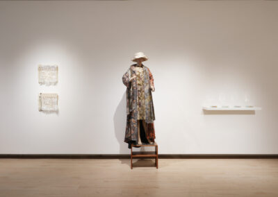 A mannequin dressed in a patterned, layered outfit and hat stands on a wooden chair before a white wall. Textile Art of ReUse hangs on the left; to the right is a shelf with glass objects. The scene is set in a gallery space.