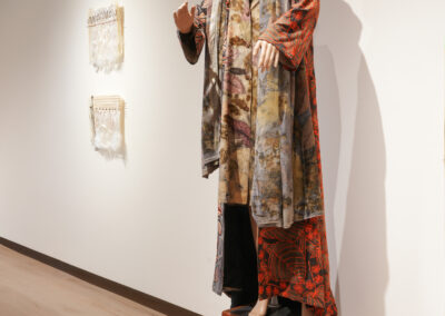 A mannequin in layered, patterned clothing and a wide-brimmed hat—showcasing the Art of ReUse—stands on a small wooden step stool in an art gallery. Upcycled textile art pieces are displayed on the white wall behind it.