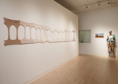 A gallery space with a long, stretched fabric art piece featuring oval cutouts—a striking example of the Art of ReUse—on a white wall, alongside a mannequin in colorful clothing and a small landscape painting in the background.