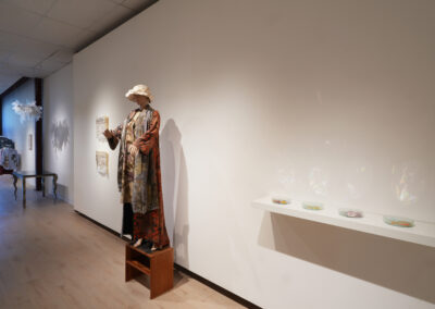 A mannequin dressed in layered, patterned clothing and a hat stands on a wooden box in a gallery space, reflecting the Art of ReUse. Artworks hang on the white wall, and small round objects are displayed on a shelf with light reflections.