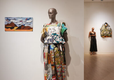 A mannequin in a colorful, patterned dress stands near a white wall displaying a small abstract painting. In the background, another mannequin and a vibrant jacket embody the gallery’s celebration of Sustainable Art and creative Reuse Art.
