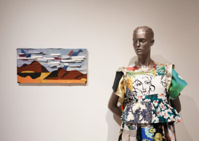 A mannequin dressed in a colorful, patterned dress stands beside a small abstract landscape on a white gallery wall, reflecting the principles of Sustainable Art and creative reuse.