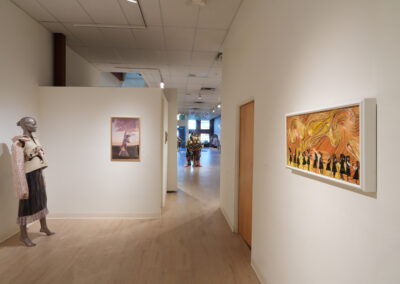 A museum hallway displays the Art of ReUse: a mannequin in upcycled clothing on the left, framed photos on white walls, and a large sculpture visible in the distance under bright lighting.