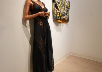 A mannequin with a copper-tone finish wears a sheer black lace dress and leans against a white wall. On the adjacent wall, an Art of ReUse colorful, abstract-patterned jacket hangs. The room has light wooden floors.