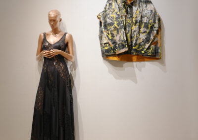 A mannequin in a long, sleeveless black lace dress stands beside a wall showcasing a colorful, abstract-patterned jacket. This stylish boutique setting highlights the Art of ReUse through unique and sustainable art fashion pieces.