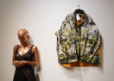 A mannequin in a black lace dress stands next to a wall where a paint-splattered, yellow and black jacket—showcasing the Art of ReUse—hangs on a hanger.