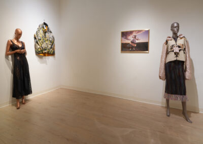 Two mannequins in stylish outfits stand in a gallery-like space; one wears a black dress, and the other sports a textured jacket and skirt. The display celebrates the Art of ReUse, with sustainable art showcased on crisp white walls.