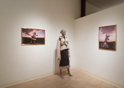 A mannequin dressed in layered clothing stands in a gallery corner, exemplifying sustainable art. It is flanked by two framed photos of a person outdoors in similar attire beneath a dramatic sky. The pale, minimalistic gallery walls and floor set the scene.