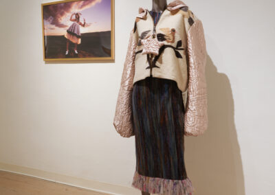 A mannequin dressed in a textured jacket with quilted pink sleeves and a multicolored skirt stands in a gallery, celebrating the Art of ReUse beside a framed photograph of a person in similar reuse art, displayed on a plain wall over a wooden floor.